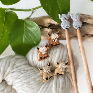 May include: Five colorful Highland cow stitch markers, four brown and one gray, on a wooden knitting needle and a ball of white yarn.