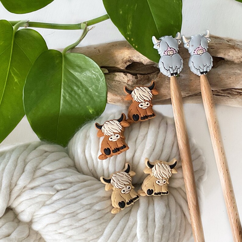 May include: Five colorful Highland cow stitch markers, four brown and one gray, on a wooden knitting needle and a ball of white yarn.
