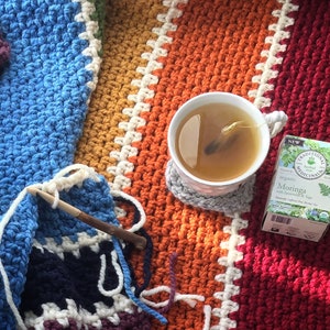 May include: A cup of tea with a tea bag in it sits on a gray coaster on top of a colorful crocheted blanket. A crocheting needle and yarn are in the lower left corner of the image. A box of Moringa tea bags is in the upper right corner of the image. The box is white with green text and a picture of Moringa leaves.