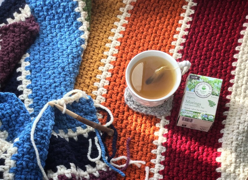 May include: A cup of tea with a tea bag in it sits on a gray coaster on top of a colorful crocheted blanket. A crocheting needle and yarn are in the lower left corner of the image. A box of Moringa tea bags is in the upper right corner of the image. The box is white with green text and a picture of Moringa leaves.