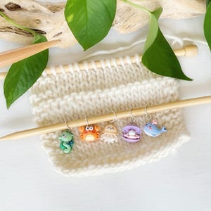 May include: A set of five colourful sea creature stitch markers, including a seahorse, crab, jellyfish, clam, and whale, are attached to a wooden knitting needle. The stitch markers are made of polymer clay and are perfect for marking your knitting projects.