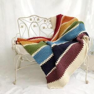 May include: A rainbow striped crochet blanket draped over a white metal bench. The blanket is made with different shades of yarn, including red, orange, yellow, green, blue, indigo, and violet.