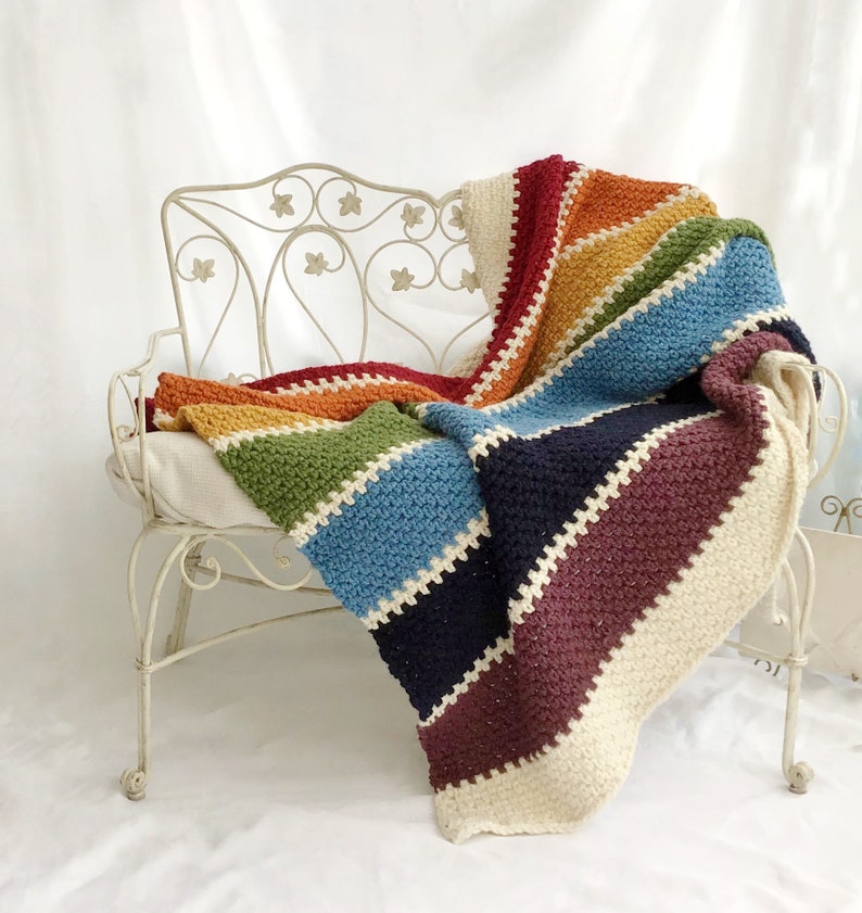 May include: A rainbow striped crochet blanket draped over a white metal bench. The blanket is made with different shades of yarn, including red, orange, yellow, green, blue, indigo, and violet.