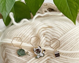 May include: Three gold metal stitch markers with different charms. The charms are a ball of yarn, a cat, and a cup of coffee with the word "coffee" on it. The stitch markers are on a white yarn background.