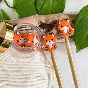 May include: Orange fox-shaped embellishments with black and white details, some inside a clear jar with a gold lid. Two fox embellishments are attached to wooden sticks. The scene includes greenery and a white yarn ball.