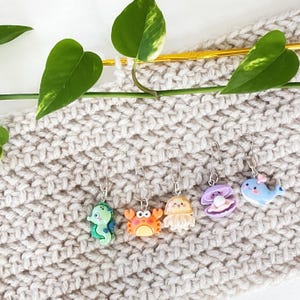 May include: A set of six colourful sea creature stitch markers. The markers are made of plastic and feature a variety of sea creatures, including a crab, a jellyfish, a clam, a whale, and a seahorse. The markers are attached to silver rings.
