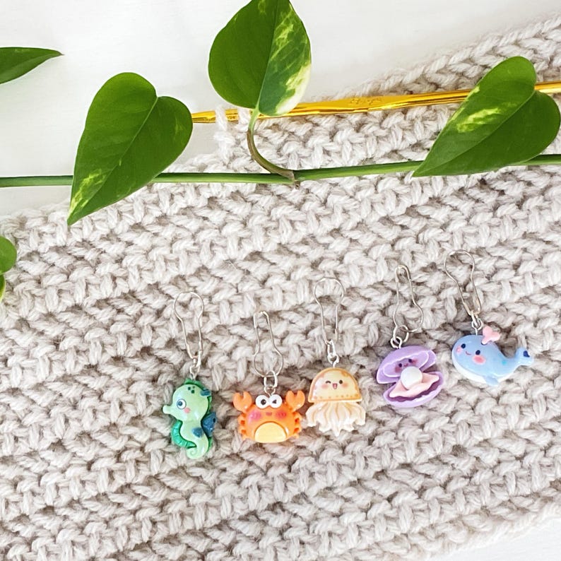 May include: A set of six colourful sea creature stitch markers. The markers are made of plastic and feature a variety of sea creatures, including a crab, a jellyfish, a clam, a whale, and a seahorse. The markers are attached to silver rings.