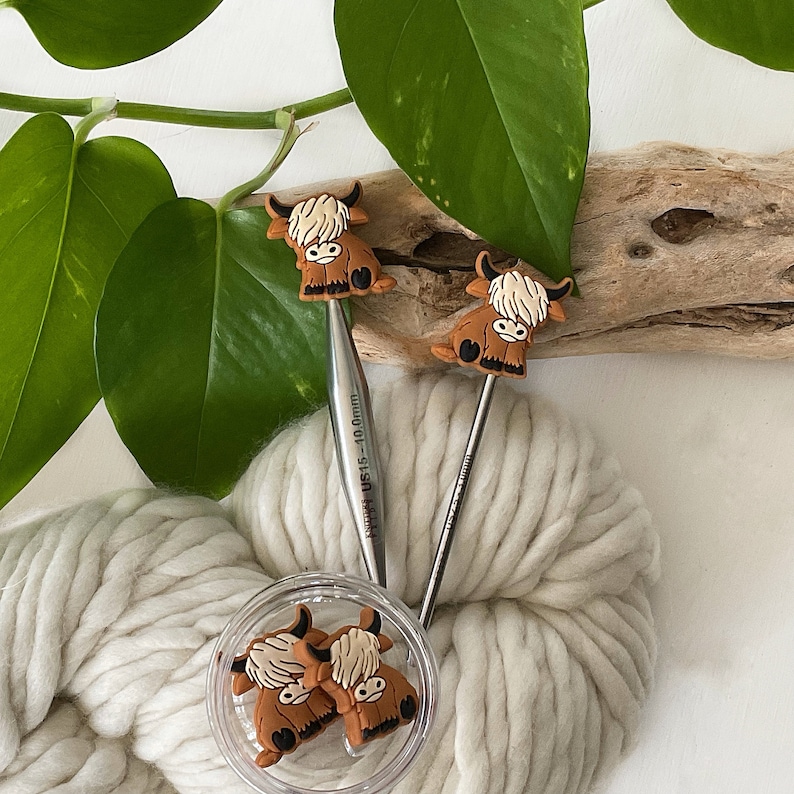 May include: Two knitting needles with brown highland cow toppers. The needles are silver and have size markings. The needles are on top of a ball of white yarn.