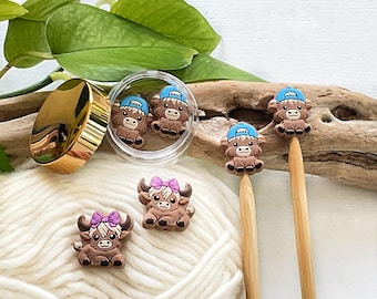 May include: A collection of Highland cow-themed craft supplies. The image features several brown cow charms with blue caps and purple bows, along with wooden knitting needles and a gold-lidded jar. The items are arranged on a white yarn background.