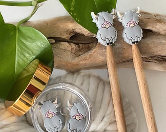 May include: Two wooden knitting needles with gray Highland cow charms attached. The needles are resting on a piece of driftwood and a white yarn ball.