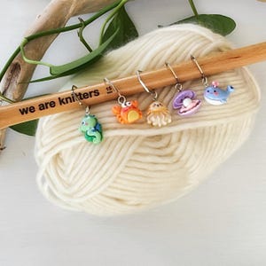 May include: A set of five colourful sea creature stitch markers on a wooden knitting needle. The markers feature a seahorse, crab, jellyfish, clam, and whale. The needle is labelled "we are knitters".