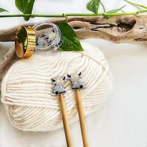 May include: Two knitting needles featuring small, grey and cream-colored highland cow charms at the top. The needles are made of light-colored wood. A small, clear plastic container with a gold lid holds additional matching cow charms.