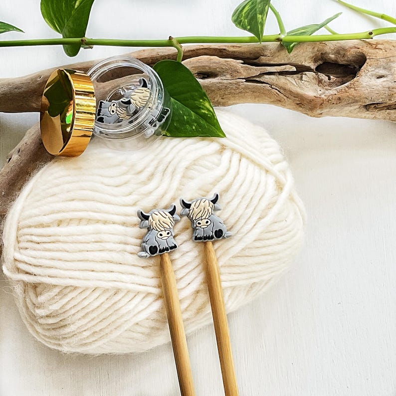 May include: Two knitting needles featuring small, grey and cream-colored highland cow charms at the top. The needles are made of light-colored wood. A small, clear plastic container with a gold lid holds additional matching cow charms.