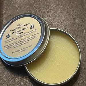 May include: Open, silver-colored tin of The Calendula Shoppe Willow Bark Salve. The lid is partially open, revealing a yellow, balm-like substance. Text on the lid describes ingredients and uses, including white willow bark and beeswax. Handmade in Jamestown, Ohio.