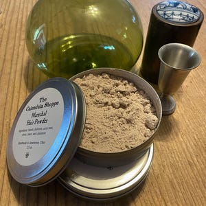 May include: A silver tin with a white label that reads "The Calendula Shoppe Marechal Hair Powder" and lists the ingredients: "Starch, dolomite, orris root, clove, mace, and cinnamon". The label also states "Handmade in Jamestown, Ohio 2.5 oz". The tin is open and contains a light brown powder.