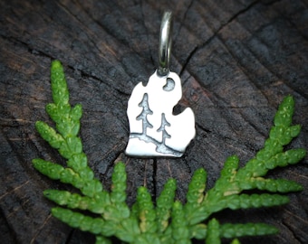 May include: A silver charm shaped like the state of Michigan, featuring a silhouette of three pine trees and a crescent moon.