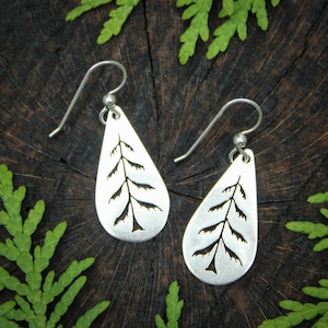 May include: A pair of silver earrings with a teardrop shape and a cut-out design of a fern leaf. The earrings are on a wooden surface.