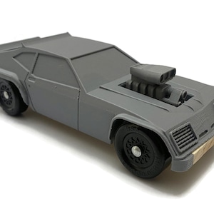 May include: A grey toy car with a large engine sticking out of the bonnet. The car has black wheels and a wooden base.