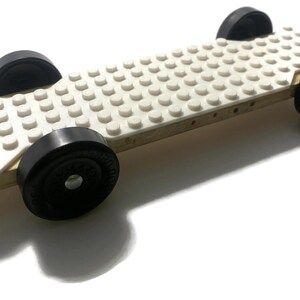 May include: A handmade toy car with a white Lego brick top and four black wheels. The car is constructed on a wooden base. The wheels are approximately 2 inches in diameter. The toy is designed for play.
