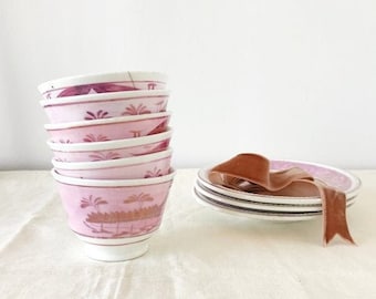 May include: A stack of six pink and white porcelain teacups with a floral design. The cups are stacked on a white surface. A pink and white saucer with a brown ribbon is in the background.