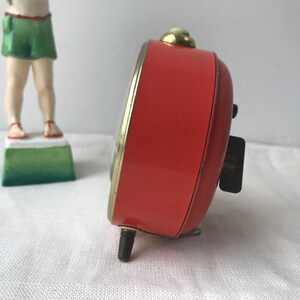 May include: A red vintage alarm clock with a gold trim and black hands. The clock is on a white surface.