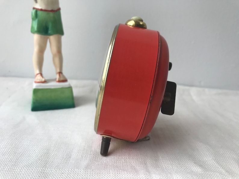 May include: A red vintage alarm clock with a gold trim and black hands. The clock is on a white surface.