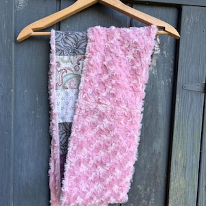 May include: A pink, rose-textured scarf with a patchwork design. The patchwork features gray, white, and purple paisley and floral patterns. The scarf is hanging on a wooden hanger against a weathered, gray-blue wooden door.