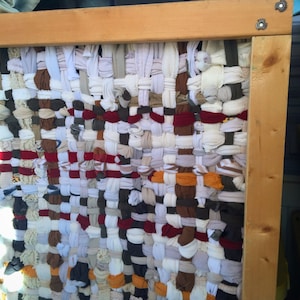 May include: A woven textile art piece with a wooden frame. The woven material is made of various fabric scraps in shades of white, brown, red, and orange. The frame is made of light-colored wood, and the textile is tightly woven.