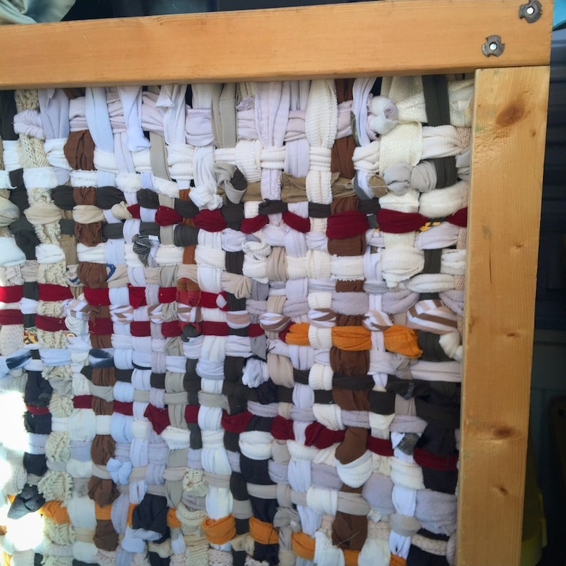 May include: A woven textile art piece with a wooden frame. The woven material is made of various fabric scraps in shades of white, brown, red, and orange. The frame is made of light-colored wood, and the textile is tightly woven.