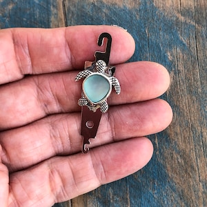 May include: A silver turtle charm with a light blue glass center. The charm is attached to a silver loop and has a silver hook on the bottom.