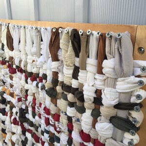 May include: A woven textile art piece made from repurposed fabric strips in various colors, including white, brown, red, and olive green. The fabric is attached to a wooden frame with screws, creating a textured, handmade wall hanging.