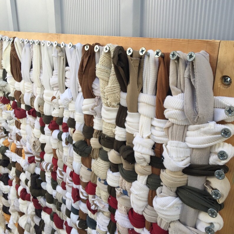 May include: A woven textile art piece made from repurposed fabric strips in various colors, including white, brown, red, and olive green. The fabric is attached to a wooden frame with screws, creating a textured, handmade wall hanging.