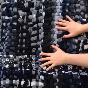 May include: A handmade rug composed of woven fabric strips in shades of black, gray, and white. The rug's texture is visible, with a mix of dark and light tones creating a patterned effect. The rug appears to be made from recycled materials.
