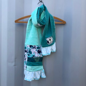 May include: A teal blue and green patchwork scarf with a white ruffle trim and a small fabric tag with a tree design.