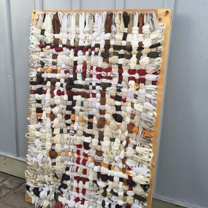 May include: A rectangular woven wall hanging with a wooden frame. The textile is made of strips of fabric in various colors, including white, brown, red, and beige. The woven pattern creates a textured surface, suitable for home decor.