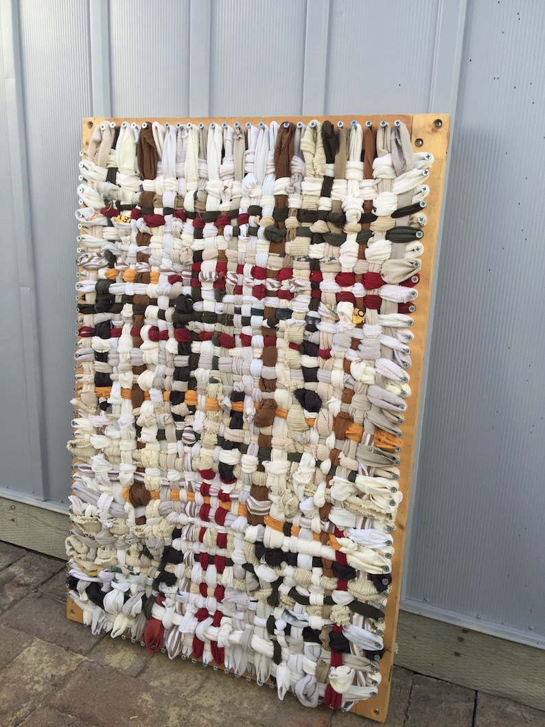 May include: A rectangular woven wall hanging with a wooden frame. The textile is made of strips of fabric in various colors, including white, brown, red, and beige. The woven pattern creates a textured surface, suitable for home decor.