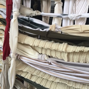 May include: Close-up of a woven textile art piece. The piece is made from various fabrics in shades of cream, beige, olive green, and burgundy. Knotted strips of fabric create a textured, layered effect, showcasing a mix of knit and woven materials.