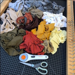 May include: A collection of colorful fabric scraps in shades of red, yellow, green, brown, and gray. A white rotary cutter with an orange blade and a pair of gray and white scissors are on a black cutting mat. A wooden loom is on the right.