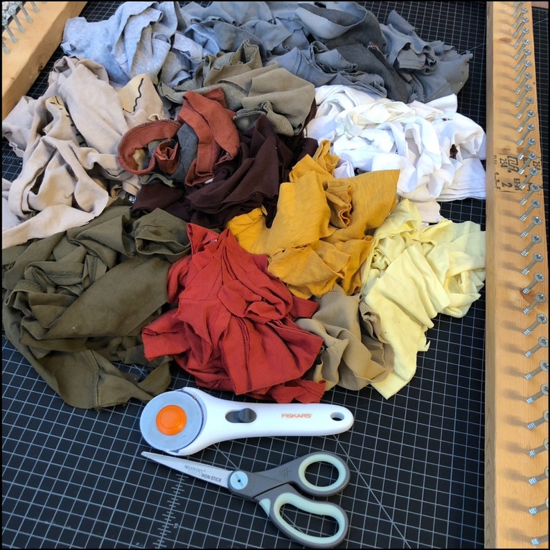 May include: A collection of colorful fabric scraps in shades of red, yellow, green, brown, and gray. A white rotary cutter with an orange blade and a pair of gray and white scissors are on a black cutting mat. A wooden loom is on the right.