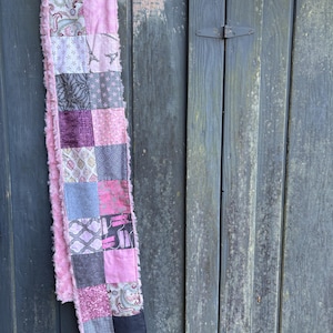May include: A patchwork scarf with a variety of pink, gray, and black patterned squares. The scarf has a soft, pink, fringed border and is hanging on a wooden hanger against a weathered wooden door.
