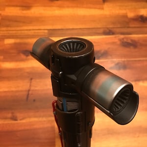 Puede incluir: Un sable de luz negro y plateado con un diseño de guarda cruzada. El sable de luz tiene un mango cilíndrico y tres hojas anguladas. Las hojas tienen una apariencia metálica desgastada con toques de azul y marrón. El mango presenta cableado rojo y azul.