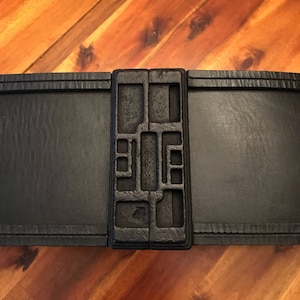 Puede incluir: Un cinturón negro con una hebilla rectangular decorativa. La hebilla tiene un diseño geométrico con cuadrados y rectángulos empotrados. El cinturón parece estar hecho de cuero o un material similar, con una superficie texturizada.