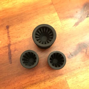 Puede incluir: Tres objetos circulares negros con un diseño estriado en forma de abanico en el interior. Los objetos son de diferentes tamaños, uno más grande y dos más pequeños. Están dispuestos sobre una superficie de madera.