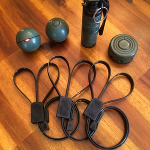 May include: An assortment of military-style props, including a green grenade, two spherical objects, a circular object, and several black loops. The items are arranged on a wooden surface, showcasing a tactical or action-oriented theme.