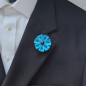 May include: A blue flower lapel pin with a black center. The pin is worn on a black suit jacket.