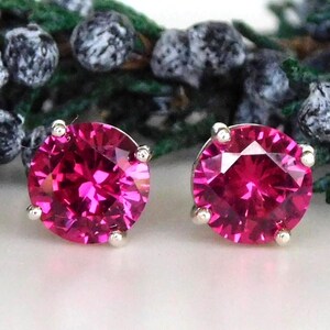May include: A pair of round-cut, vibrant fuchsia-pink stud earrings set in silver-toned metal prongs. Each earring features a faceted gemstone that reflects light, showcasing a brilliant, deep pink color and a classic circular shape.