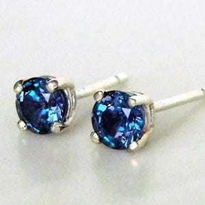 May include: A pair of stud earrings featuring round, deep blue gemstones set in silver-colored metal. The gemstones have a faceted appearance, reflecting light. The earrings are designed with a classic, minimalist style.
