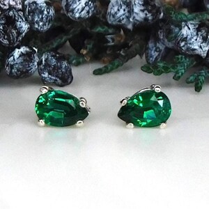 May include: A pair of stud earrings featuring pear-shaped, faceted emerald green gemstones set in silver-toned metal prongs. The earrings are positioned side-by-side, showcasing the vibrant green color and the clear, reflective quality of the stones.
