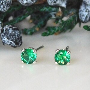 May include: A pair of stud earrings featuring round, faceted, bright green gemstones set in silver-colored metal. The earrings are displayed on a white surface with green and gray foliage in the background.
