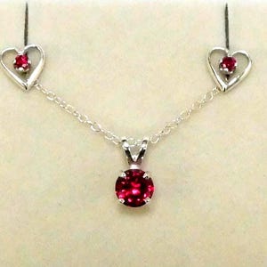 May include: A silver necklace set featuring a central round, faceted red gemstone pendant suspended from a delicate chain, flanked by two matching silver heart-shaped stud earrings, each containing a small, round, faceted red gemstone.